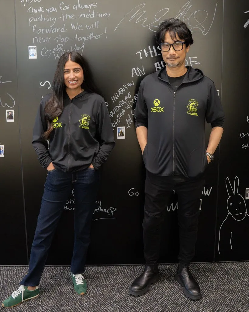 Asha Sharma e Hideo Kojima em pé lado a lado vestindo casacos pretos com os logos do Xbox e da Kojima Productions, em frente a uma parede escura repleta de autógrafos.