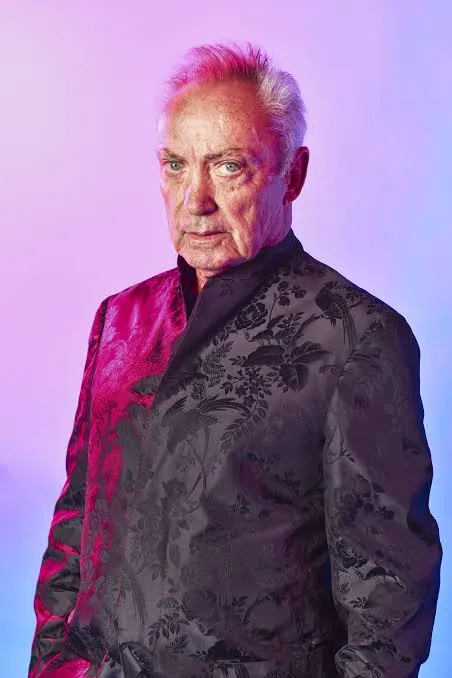 Retrato do ator veterano Udo Kier sob iluminação neon em tons de roxo e rosa, vestindo um paletó escuro com estampas florais. O ator integra o elenco principal do jogo OD Knock de Hideo Kojima.